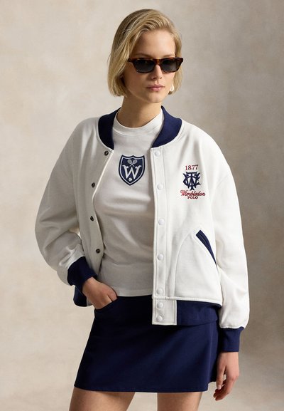Woman wearing sunglasses, white varsity jacket with navy trim and logos, white t-shirt, and navy skirt, posing with one hand in pocket.