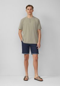 Licht olijfgroene T-shirt met korte mouwen en henley-stijl met drie knopen, gecombineerd met donkerblauwe denimshorts en bruine sandalen. Vlakke textuur en opgerolde zomen.