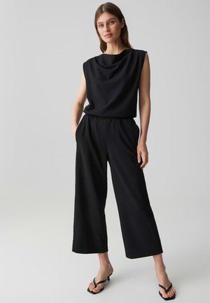 Frau steht mit Händen in den Taschen, trägt ärmelloses schwarzes Oberteil, weite schwarze Hosen und schwarze Sandalen mit Absatz vor grauem Hintergrund.