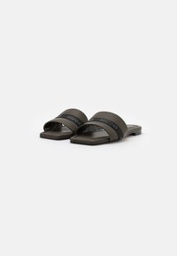 Une paire de slides plates de couleur vert olive avec des larges sangles, intégrant des bandes noires et une marque en relief "HUGO" sur fond blanc.
