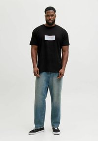 Camiseta de algodón negra con un logotipo rectangular en rojo y blanco, combinada con unos vaqueros de denim azul claro de corte holgado y zapatillas negras.