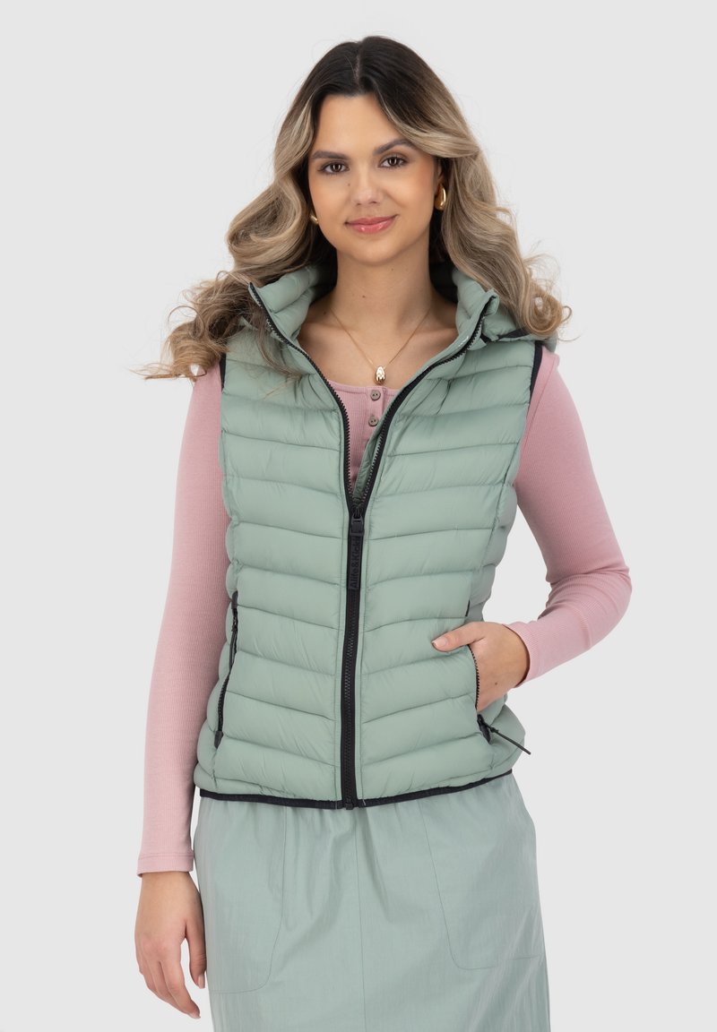 Gilet imbottito verde chiaro con colletto alto, zip frontale e tasche laterali. Indossato sopra una maglietta a maniche lunghe rosa, con cuciture orizzontali.