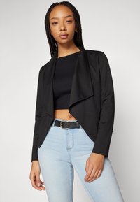 Svart drapert blazer med en myk tekstur, åpen front og lange ermer, kombinert med en svart kort topp og lyseblå jeans.