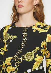 Vestido negro con un vibrante patrón floral amarillo y acentos de cadena dorada. Escote redondo con el texto "JEANS COUTURE" integrado en el diseño.