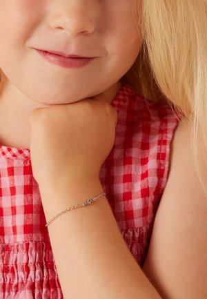 Bracelet en argent avec des accents de pierres colorées ; design de chaîne délicat ; porté sur le poignet d'un enfant ; le fond est un motif vichy rouge.
