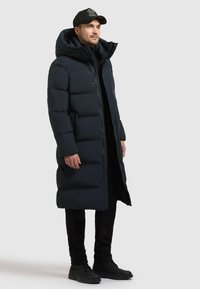 Tume sinine polsterdatud pikk jakk kõrge kraega ja kapuutsiga, millel on nupud ja lukud. Kandel mustade pükste ja jalatsitega.