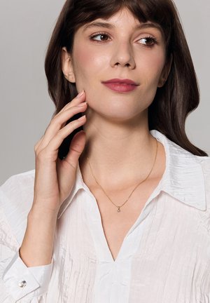 Złoty naszyjnik z małym wisiorkiem, noszony na białej, teksturowanej koszuli. Ręka modelki delikatnie dotyka jej szyi.