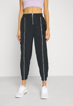 Kvinde model iført sorte bukser med vertikale lynlåse foran og en maveblottende lavendel-farvet, blød crop top, kombineret med hvide sneakers.