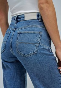 Acercamiento de una persona que lleva puestos unos pantalones de mezclilla azul de cintura alta con bolsillos traseros y una etiqueta de marca de cuero blanco, combinados con una blusa sin mangas blanca.