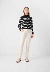 Čierno-biely pruhovaný cropped kardigán so zlatými gombíkmi, kombinovaný s krémovými priliehavými nohavicami a svetlými topánkami s dekoračnými detailmi.