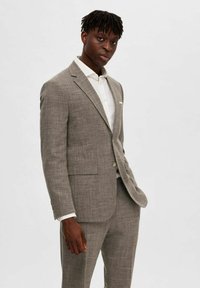 Costume gris clair en tissu texturé. Comprend un blazer à bouton unique, des revers en notch et un pantalon ajusté. Porté sur une chemise blanche.