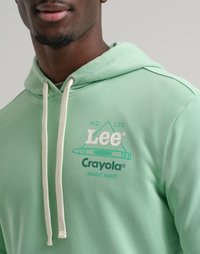 Primo piano di una persona che indossa un hoodie verde menta con lacci bianchi e un logo che combina i marchi Lee e Crayola sul petto.