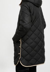 Sort quiltet dunfrakke med hætte, der har et tekstureret diamantmønster, beige kantning og trykknapdetaljer langs sidesømmen.