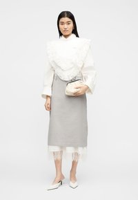 Une femme debout porte un chemisier blanc à volants en dentelle, une jupe midi grise, un ourlet en dentelle blanche, des escarpins blancs, et tient un petit sac à main beige.