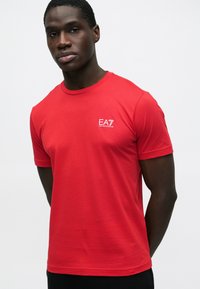 T-shirt rossa in cotone a maniche corte, con un piccolo logo bianco EA7 sul petto a sinistra. Il tessuto appare liscio e leggero.