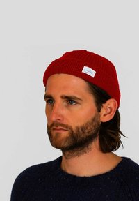 Red ribbed knit beanie with a small white label on the front. Solid color and textured fabric, worn by a person in a blue sweater.