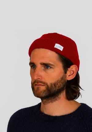 Red ribbed knit beanie with a small white label on the front. Solid color and textured fabric, worn by a person in a blue sweater.