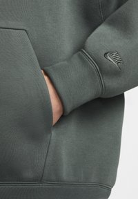 Tmavě zelená mikina Nike s texturovaným povrchem, obsahující klokaní kapsu a malé vyšívané logo na rukávu.