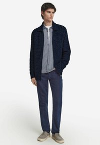 Marinblå texturerad stickad cardigan, ljusgrå knäppskjorta, mörkblå byxor och beige sneakers. Cardiganen har krage och öppen framsida.