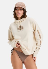 Sudadera beige con un diseño impreso en la parte frontal y texto en la manga, acompañada de un bikini de parte inferior marrón y una gorra marrón claro.