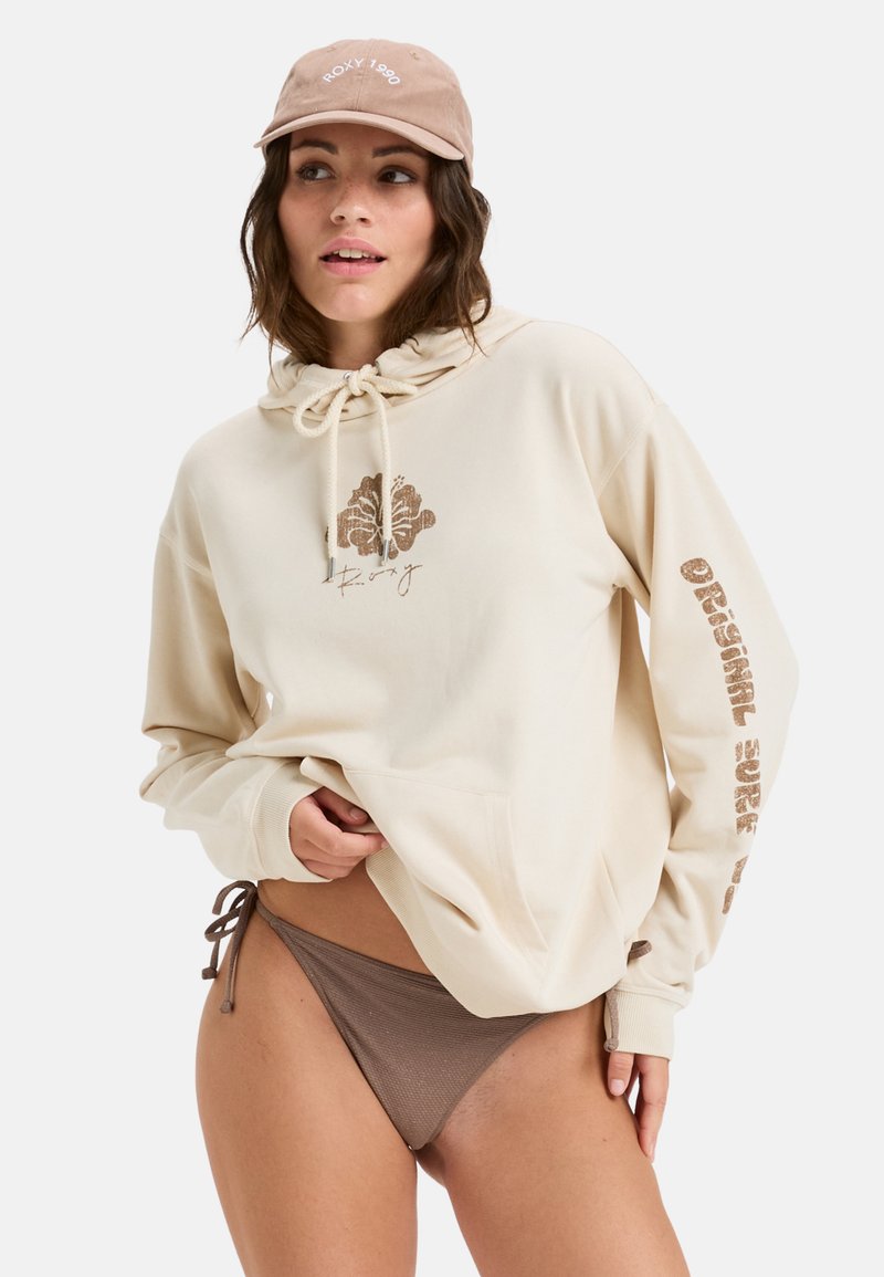 Sudadera beige con un diseño impreso en la parte frontal y texto en la manga, acompañada de un bikini de parte inferior marrón y una gorra marrón claro.