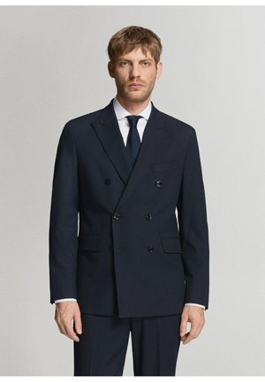 Uomo con capelli castano chiaro che indossa un abito doppiopetto scuro, camicia bianca e cravatta scura, in piedi davanti a uno sfondo chiaro e semplice.