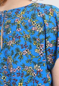 Modrý květinový top s krátkými rukávy, zdobený vzorem růžových, žlutých a zelených květin, s malým otvorem ve tvaru klíče na zadní straně.