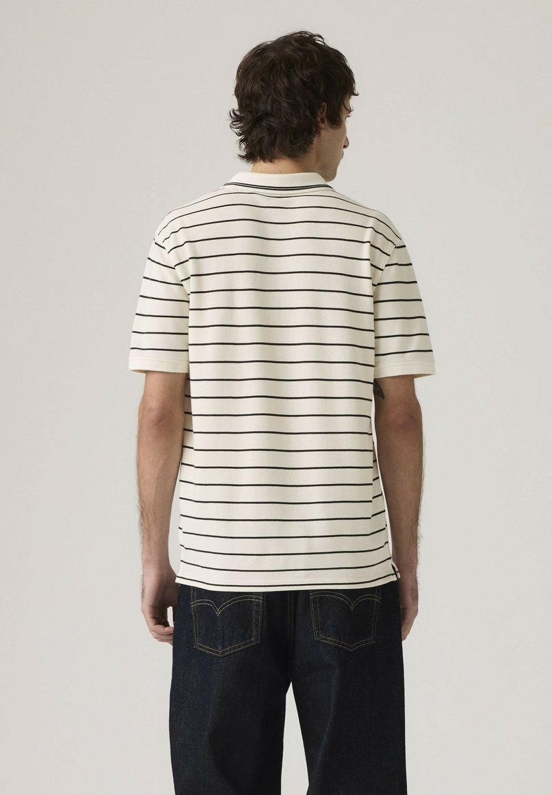 Hombre de espaldas que lleva una camisa blanca de manga corta con finas rayas horizontales negras y vaqueros azul oscuro.