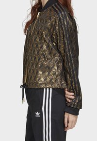 Černá bomber bunda se zlatým geometrickým vzorem, zipem vpředu, žebrovaným límcem a černými pruhy na rukávech. V kombinaci s černými sportovními kalhotami.