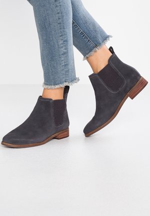 Person iført mørkegrå ruskind Chelsea-støvler med brune såler og lyseblå jeans med frynset kant på hvidt gulv.