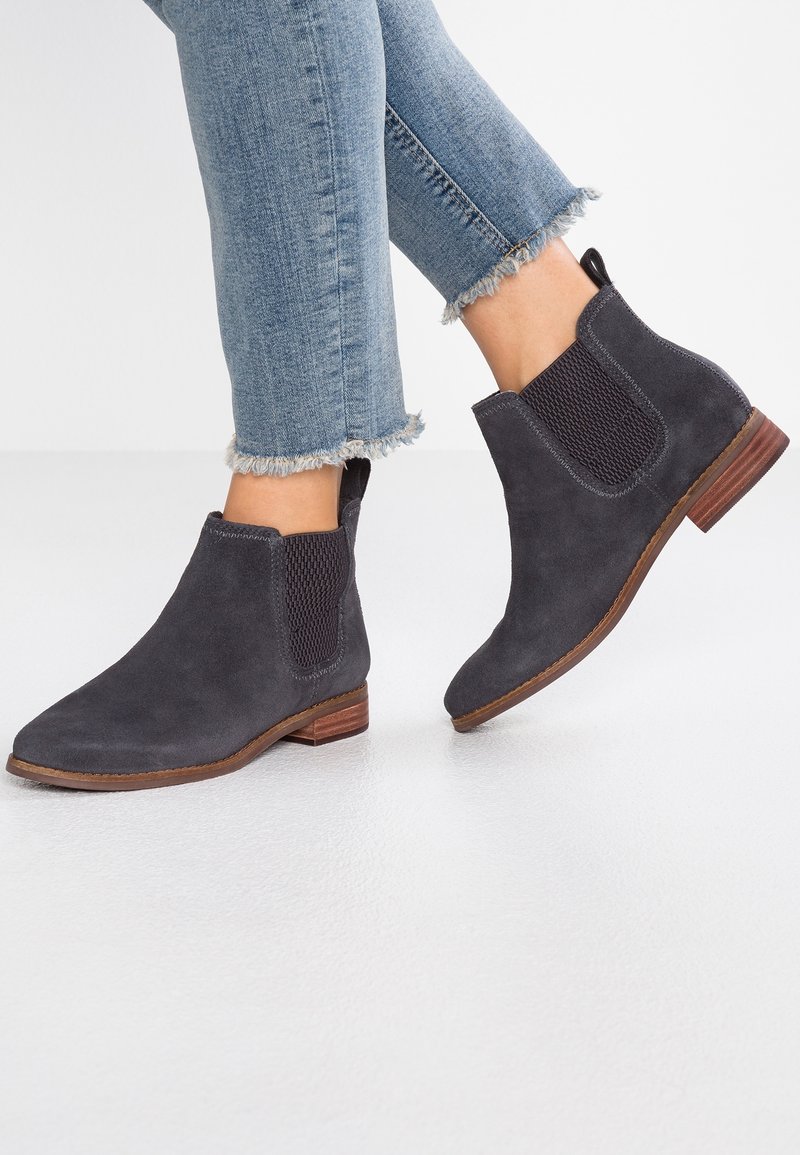 Personne portant des bottines Chelsea en daim gris foncé avec des semelles marron et un jean bleu clair à ourlet effiloché sur un sol blanc.