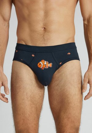 Męski tors w ciemnych slipach z pomarańczowymi wzorami ryby klauna z przodu i po bokach, stojący na jednolitym tle.