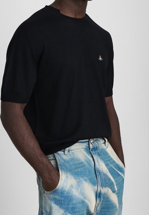 Mann trägt schwarzes kurzärmliges Strickshirt mit kleinem buntem Emblem, Hände in den Taschen, kombiniert mit hellblauen Acid-Wash-Jeans.