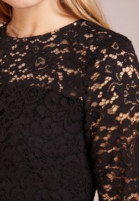 Svart spetsblus med utsökta blommönster och genomskinlig struktur. Rund halsringning och långa ärmar som betonar den detaljerade designen.