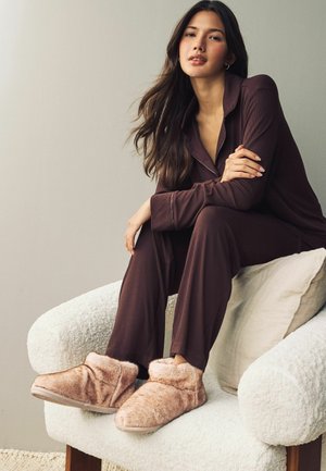 Gemütliche beigefarbene Kunstfellschuhe mit einem weichen umgeschlagenen Bund liegen auf einem weißen strukturierten Sessel und werden mit einem dunkelbraunen Schlafanzug kombiniert.