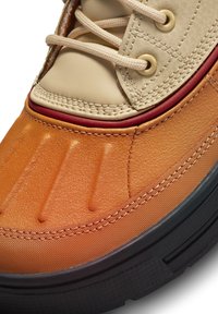 Orange and beige sneaker featuring a textured upper, lacing system, and black sole; has stitched accents and bold, shaped details.