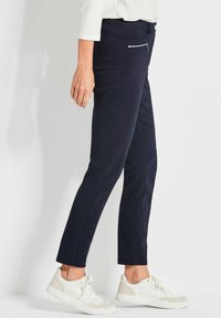 Marineblauwe broek met een slim fit, voorzien van een ritszak, zijnaad en gecombineerd met witte sneakers op een neutrale achtergrond.