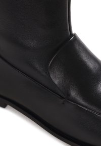 Gros plan d'une botte en cuir noir lisse avec une couture simple et un détail en cuir plié sur la partie supérieure.