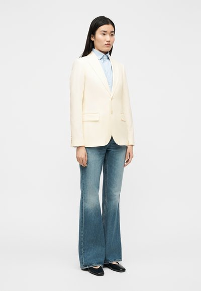 Γυναίκα με κρεμ σακάκι πάνω από ανοιχτό μπλε πουκάμισο με κουμπιά, καμπάνες μπλε τζιν και μαύρα παπούτσια με μυτερά δάχτυλα, στέκεται μπροστά σε λευκό φόντο.