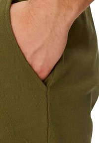 Olijfgroene stof met een gestructureerd patroon; voorzien van een zijzak en een gedeeltelijk zicht op een hand die in de zak rust.