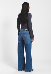 Vaqueros de mezclilla azul de cintura alta con un ajuste de pierna ancha, que presentan un diseño clásico de cinco bolsillos y una etiqueta de marca en la parte trasera, llevados con una camiseta gris ajustada.