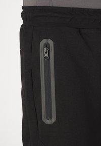 Sorte joggerbukser med en sidelomme med lynlås, struktureret stof og elastisk talje. Lynlåsen har en sort trækflap.
