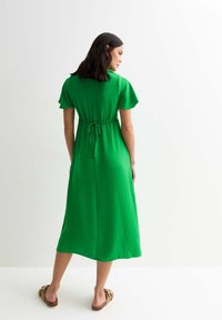 Vestido verde, hasta la rodilla, con mangas cortas, tela texturizada y detalle de cinturón atado en la cintura. La modelo lleva sandalias marrones.