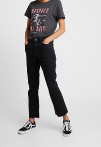 Černé džíny s roztrhaným lemem, v kombinaci s šedým tričkem s růžovým grafickým designem. Tenisky jsou černé plátěné s bílými detaily.