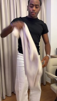 Čierna krátka rukávová košeľa, biele slim-fit nohavice, drží svetlo béžový odev. Hladká textúra, minimalistický dizajn, vnútorné prostredie s neutrálnymi tónmi.