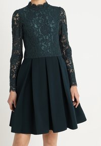 Mörkgrön klänning med en spetstopp med blommönster och långa ärmar, kombinerad med en plisserad kjol gjord av ett solidt, strukturerat tyg.