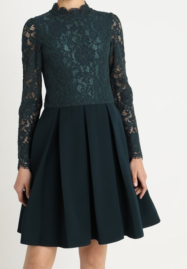 Mörkgrön klänning med en spetstopp med blommönster och långa ärmar, kombinerad med en plisserad kjol gjord av ett solidt, strukturerat tyg.