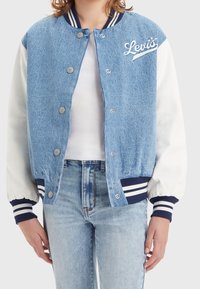 Džínová varsity bunda se světle modrým tělem, bílými rukávy, námořnickými pruhovanými ribbed manžetami, patentními knoflíky a vyšívaným logem "Levi's" na hrudi.