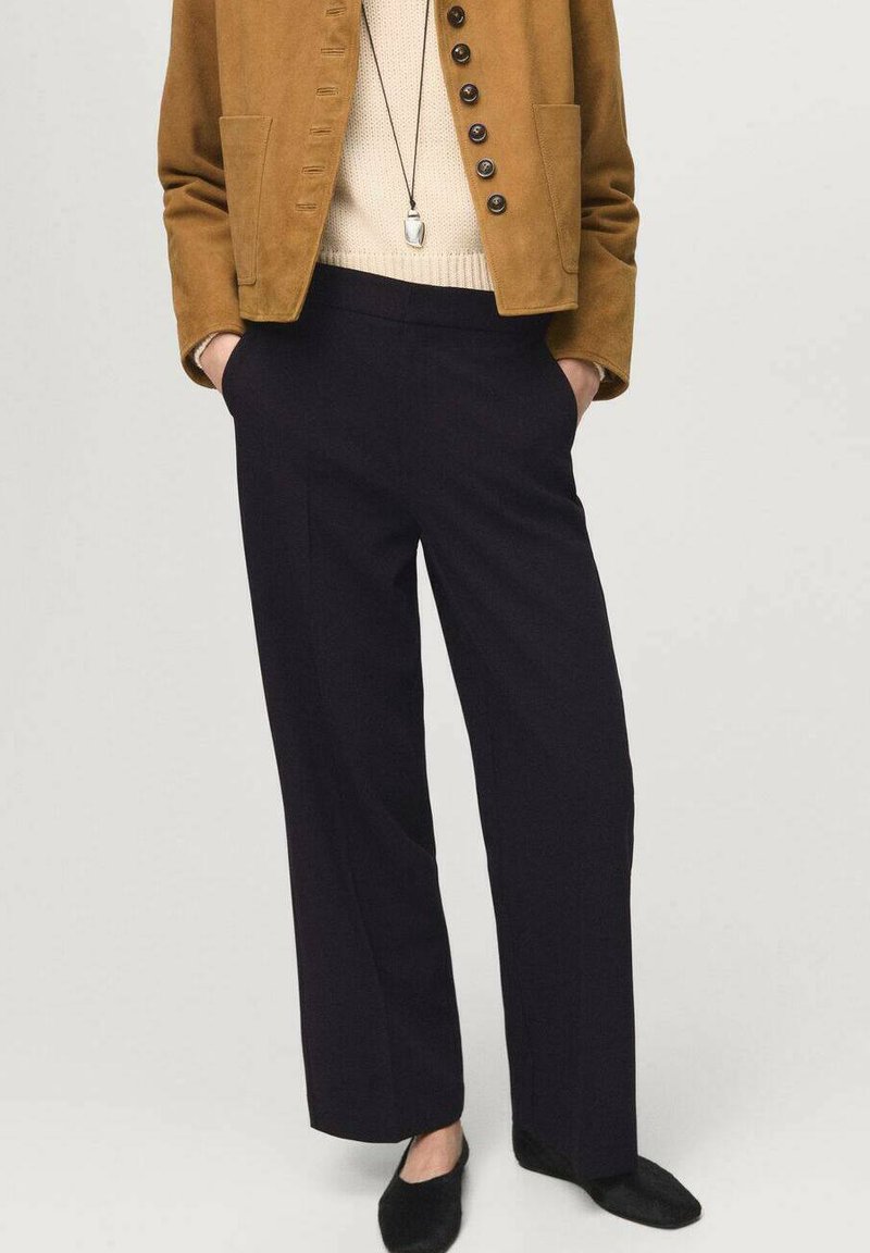 Veste en daim marron avec des accents en bouton, haut en tricot crème et pantalons larges bleu foncé. Des chaussures noires complètent la tenue.