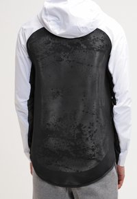 Svart och vit lättviktsjacka med meshnack, huva och rundad nederkant. Har en texturerad, mönstrad yta på det svarta materialet.